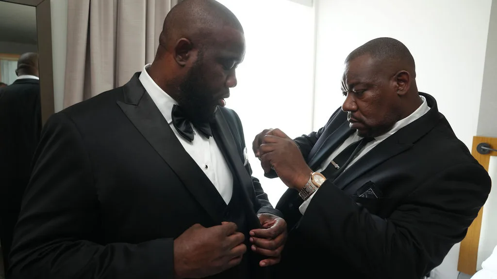 A groomsman fastens the final button on Tre's tuxedo jacket, black-tie wedding morning in New Jersey
