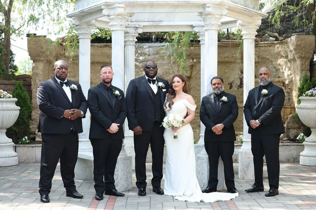 Becky with Tre's groomsmen in all-black tuxedos in front of the garden waterfall and gazebo — black-tie wedding portrait, New Jersey