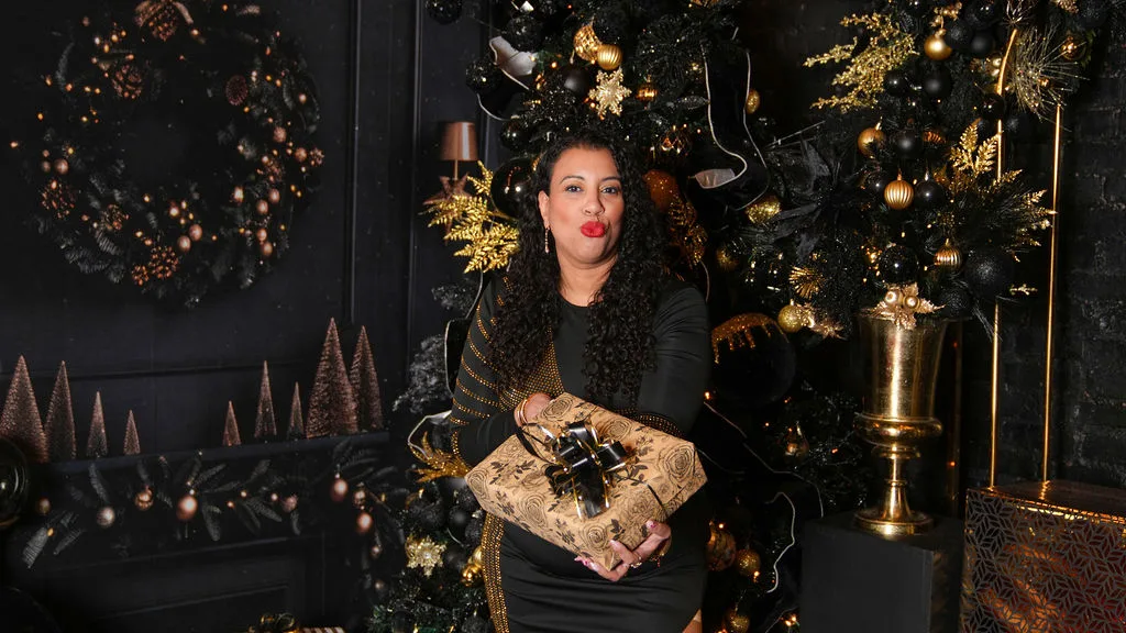 A wide frame of mom holding a wrapped gift, gold wreath and dark brick behind her — editorial Christmas portrait, Maximus Visions, New Jersey