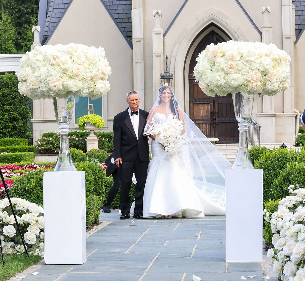 Bride and her father walk the chapel aisle between towering white rose pedestals at Park Chateau Estate, NJ