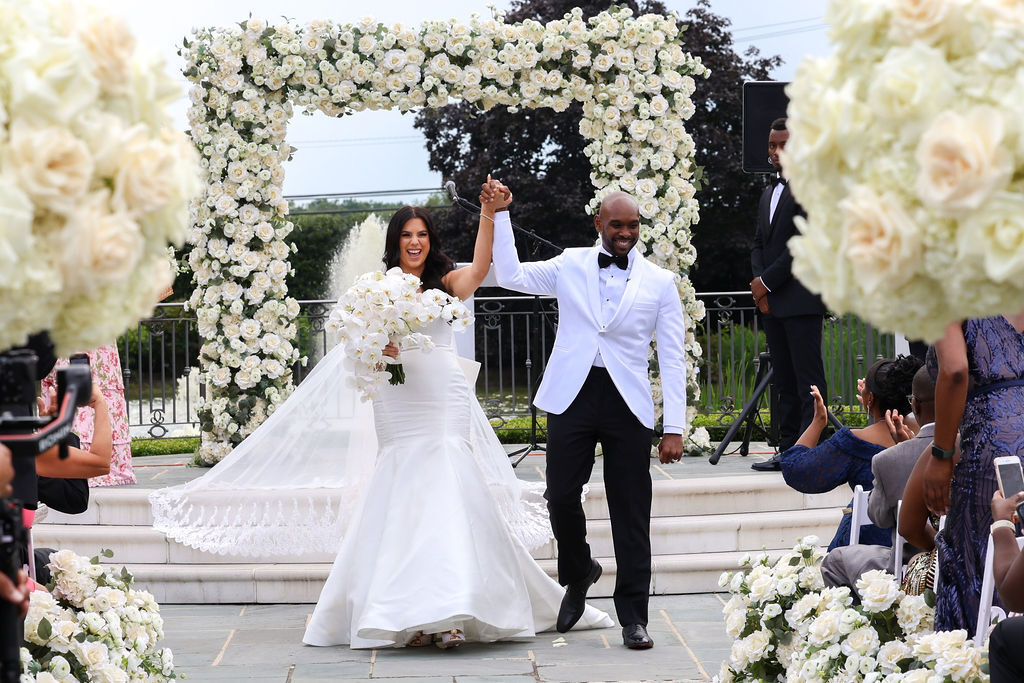 Newlyweds raise hands in celebration under a white rose ceremony arch at Park Chateau Estate, NJ