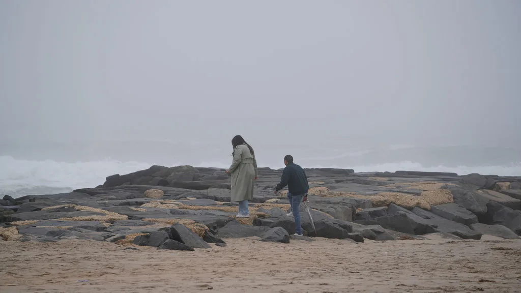 The couple climbs onto the dark jetty rocks above the surf, fog rolling behind them — New Jersey shore