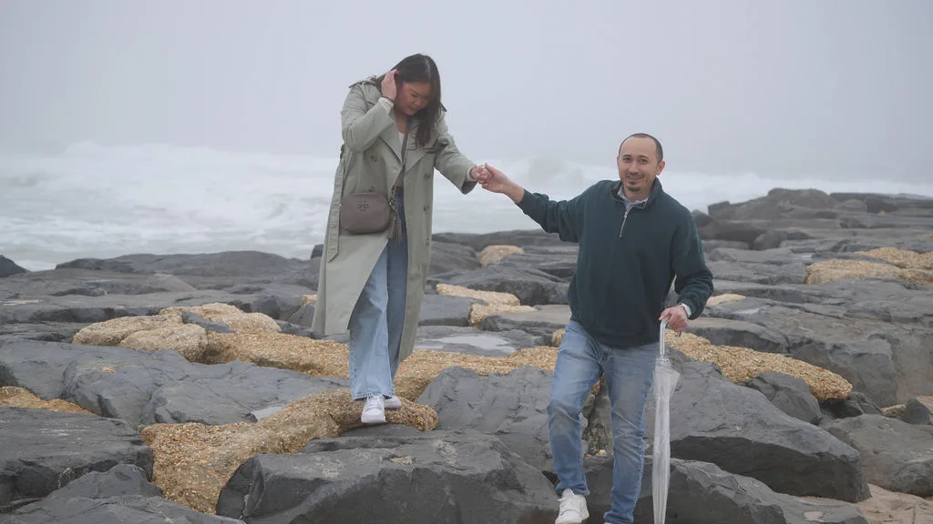 He reaches back to help her across the slick jetty rocks — candid moment before the proposal, New Jersey shore