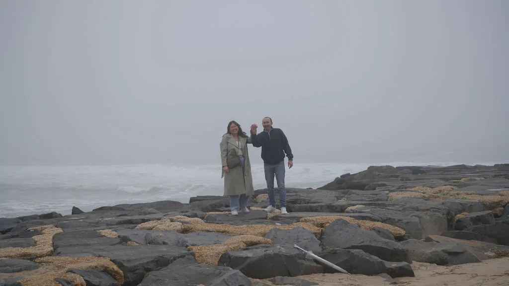 The couple stands small against the wide fog-wrapped jetty and open ocean — cinematic proposal photography by Maximus Visions, New Jersey