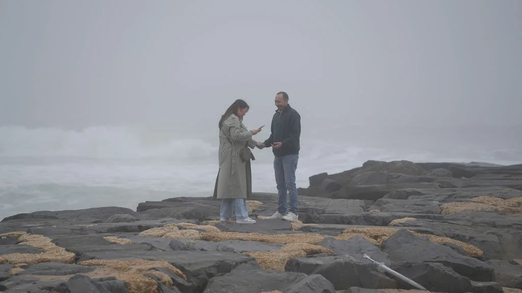 He reaches for her hand on the jetty as she looks down at her phone — the last ordinary moment before the proposal, New Jersey