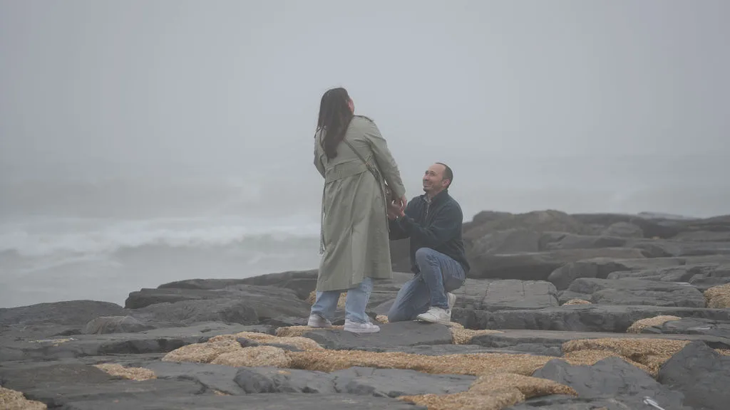He drops to one knee on the jetty rocks with the fog and ocean behind them — surprise proposal, New Jersey shore
