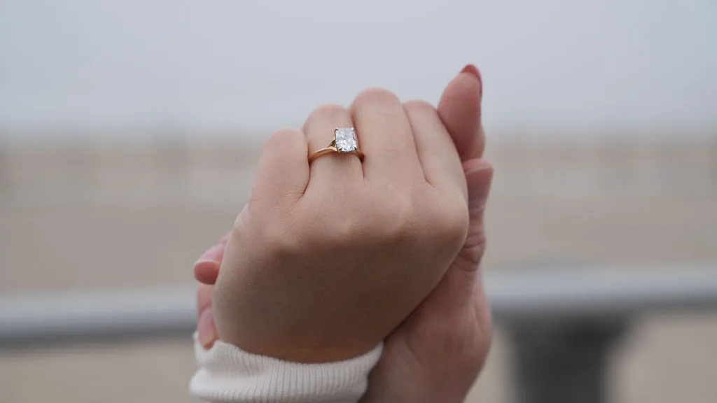 The engagement ring on her hand, fingers laced through his, the foggy shore soft behind — proposal detail photography by Maximus Visions, New Jersey